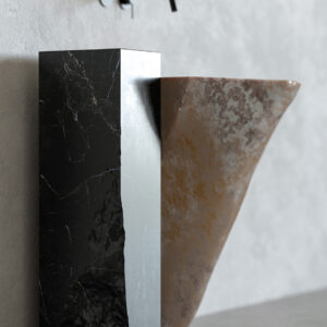 Meteora reclaimed marble sink made from a rough-cut grey stone block, paired with a dark marble counterweight block and matte-black wall-mounted tap, set against a rust-toned textured wall with marble offcuts nearby.