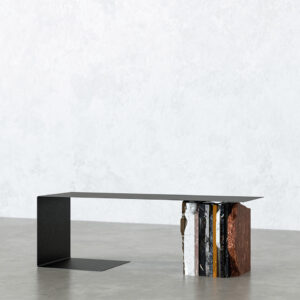Pausa bench with a thin black iron seat on an L-shaped metal leg, supported on the other end by a block of layered reclaimed marble offcuts with rough edges, photographed on a concrete floor.