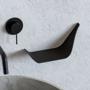 Wall-mounted Dalí shelf in matte-black metal with an upturned end, installed on a textured light wall beside a black shower control; marble basin edge in foreground.