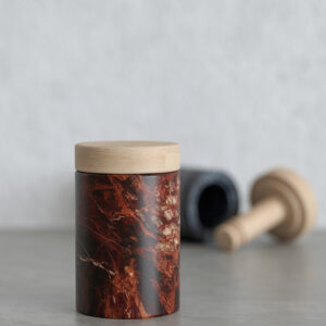 Burgundy veined marble canister with a light wood lid on a grey surface; blurred black marble cylinder and wooden dispenser parts in the background.