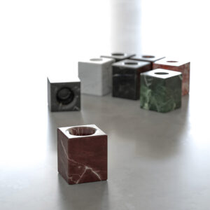Red marble cube toothbrush holder with a round top opening in focus, with other Gesti marble cube holders in white, black and green in the background on a grey surface.